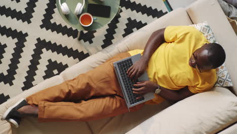 afro-american man lying on sofa and working on laptop