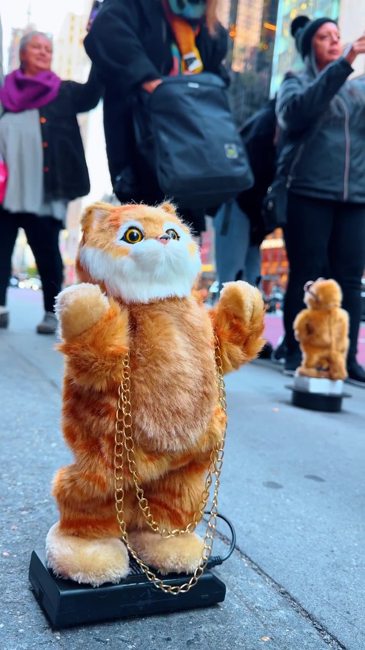 New York, AS, 5 Oktober 2025: Mainan kucing jahe animasi dengan rantai emas di trotoar. Kucing jahe mewah elektronik dengan rantai emas berdiri di atas kemasan baterai di trotoar kota.