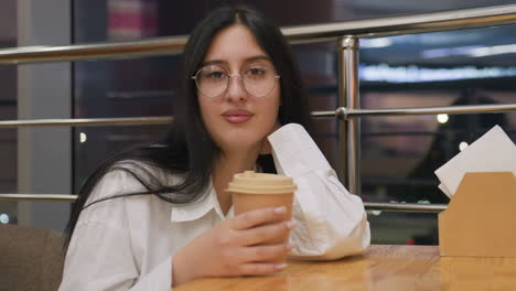 young lady leans on wooden table holding coffee cup with relaxed pose and seductive look while seated indoors near shiny railings and large glass window with blurred lights of busy city night outside