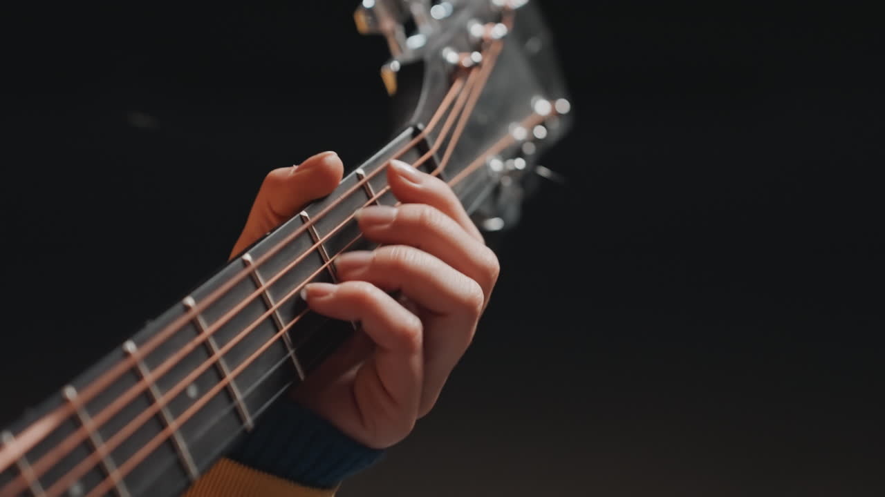 Primo piano della mano su una chitarra acustica che forma accordi aperti con una pennata lenta e controllata, con la manica di un maglione di lana visibile. La scena è avvolta da una calda luce color ambra, creando un'atmosfera di prova intima, mentre il musicista affina il ritmo e il tono.