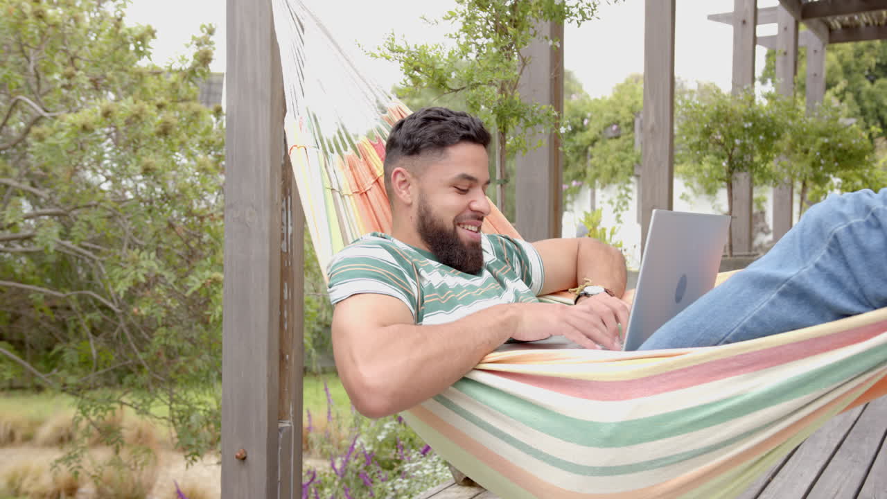 Relaxing in hammock, man using laptop and smiling in outdoor setting