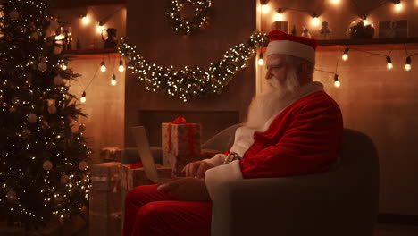 santa claus se sienta en un sofá suave en una sala de estar bellamente decorada con luces y guirnaldas. tiene una computadora portátil en el regazo y escribe texto con una mano. imágenes de alta calidad 4k
