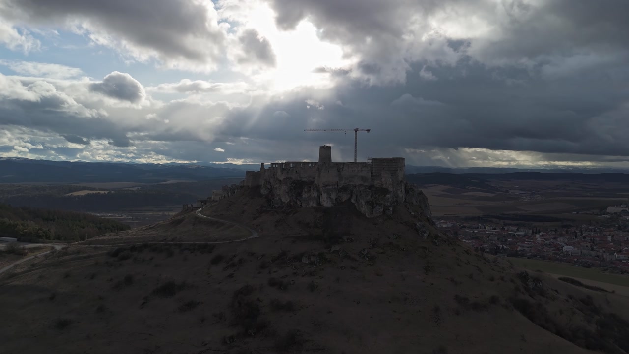 Spiš Castle, a medieval hilltop fortress in eastern Slovakia, dominates the rural landscape with its extensive stone ruins, defensive walls, and towers in this wide aerial panorama under a cloudy sky.