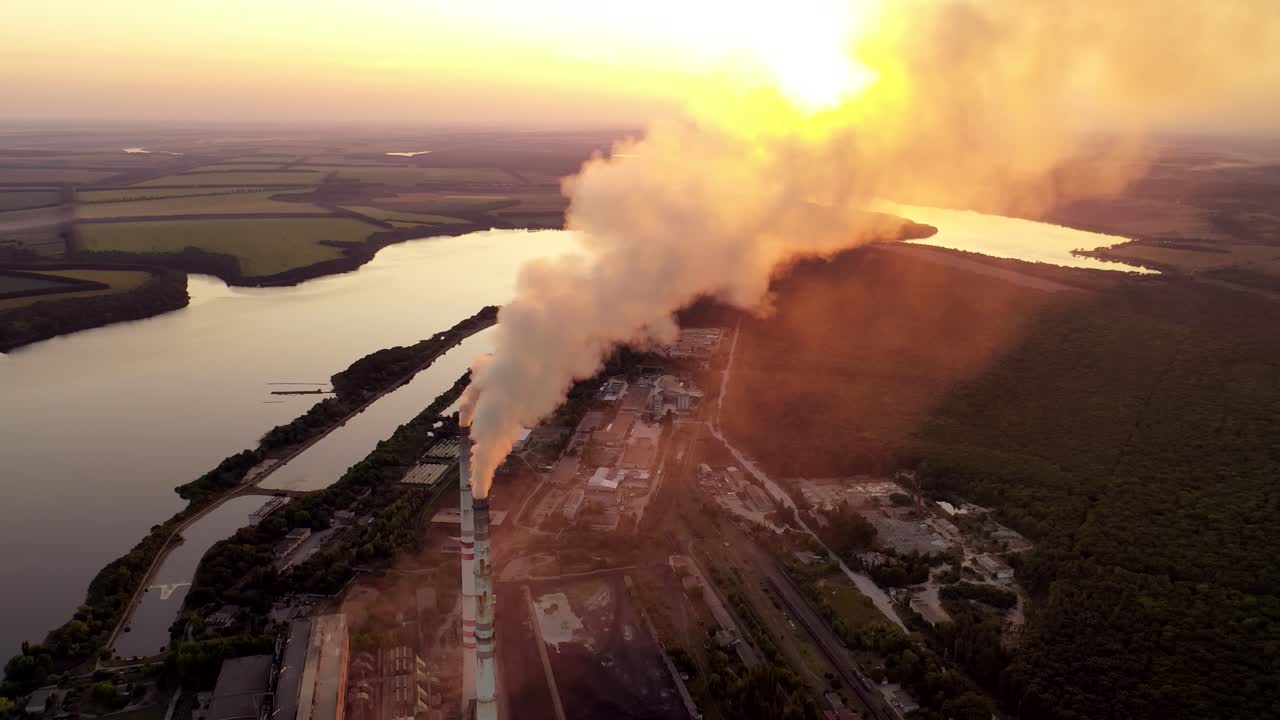 Aerial view of a factory among nature. Industrial pipes pollute the atmosphere with smoke at sunset. Beautiful river next to the huge metallurgy plant.