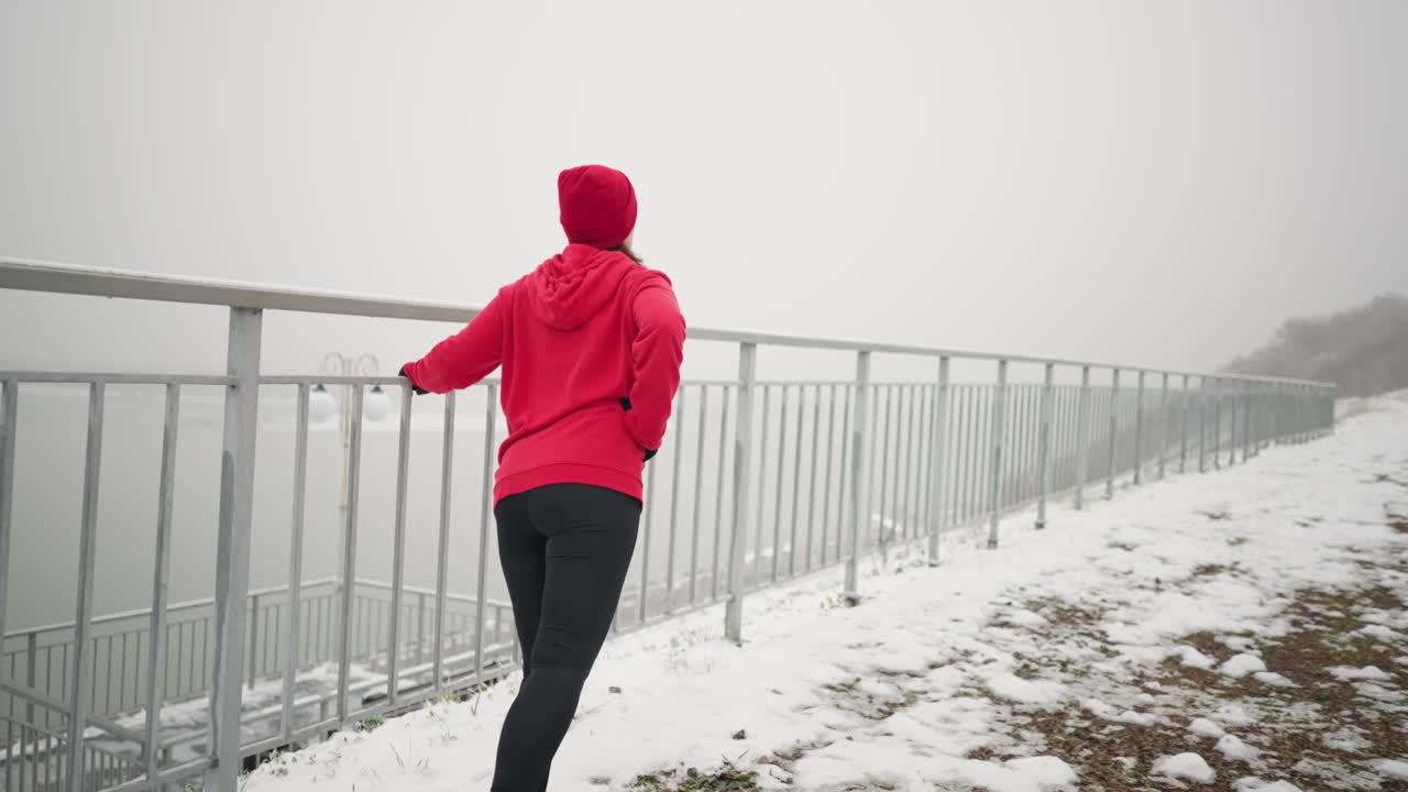 은 후디와 베니를 입은 여성이 겨울 동안 야외에서 운동을 하며, 눈 인 땅, 계단, 안개가 가득한 분위기에서 다리를 는 동안 균형을 잡기 위해 철의 레일을 들고 있습니다.