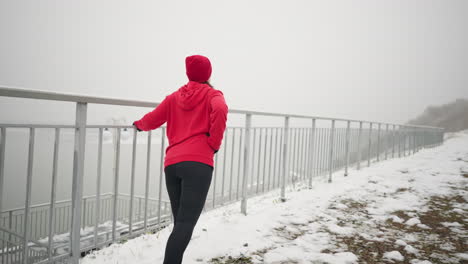 mujer con capucha roja y gorra haciendo ejercicio al aire libre durante el invierno, sosteniendo una barandilla de hierro para el equilibrio mientras estira la pierna con el suelo nevado, la escalera y la atmósfera brumosa