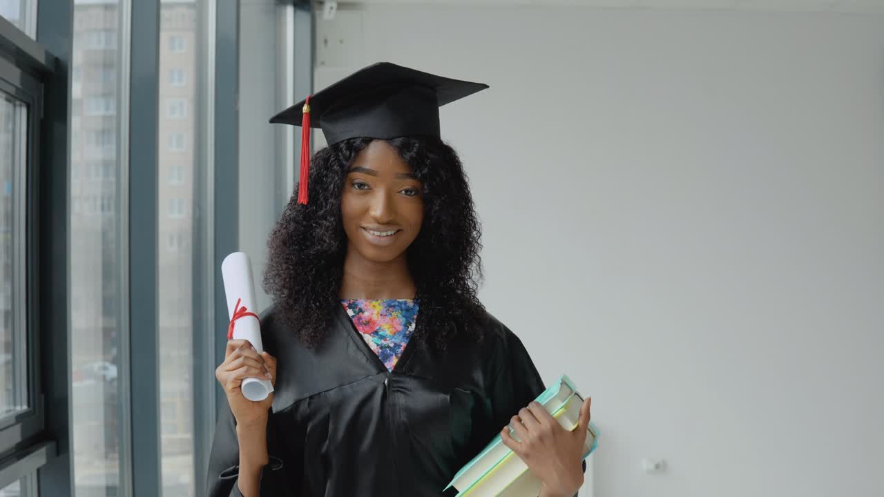 joven graduada afroamericana de pie frente a la cámara con un diploma y libros en las manos. la estudiante lleva una túnica negra y un sombrero de maestro cuadrado.