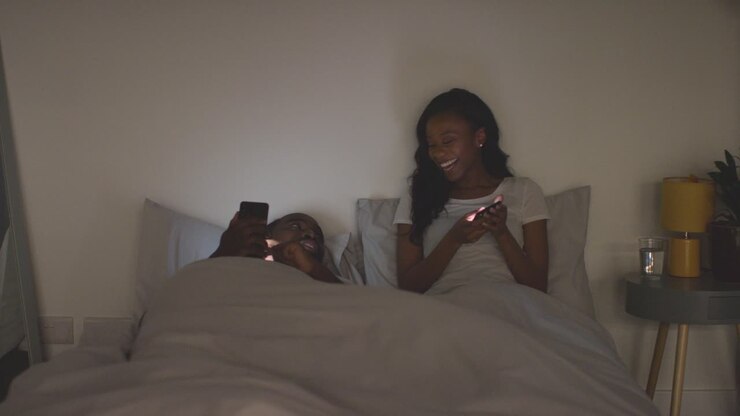 Giovane coppia che si rilassa a casa di notte a letto guardando insieme i telefoni cellulari 1