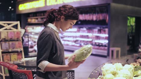 Young-beautiful-brunette-girl-in-her-20's-picking-out-napa-cabbage-and-cauliflower-and-putting-them-into-shopping-cart-at-the