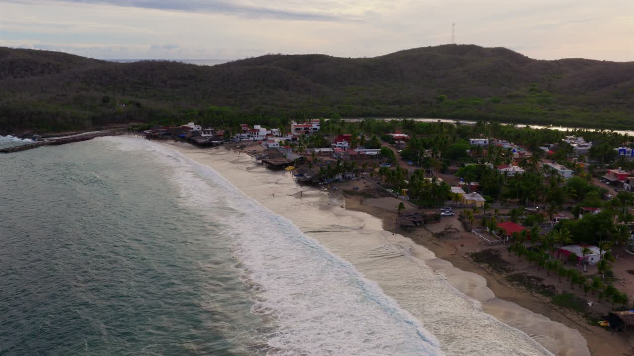 Drone video captures Punta Perula's picturesque Jalisco coastline, waves gently lapping a serene beach, vibrant homes clustered under soft, diffused evening light, lush tropical landscape