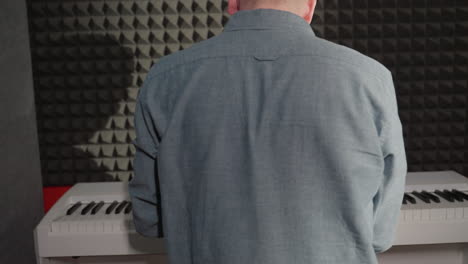 close-up back shot of a man in a blue shirt playing a white piano, with his shadow clearly visible on a textured black acoustic wall