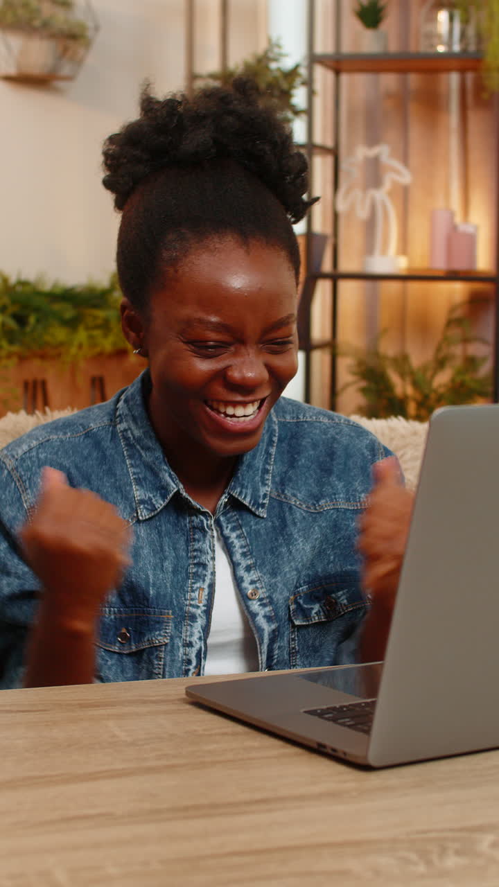 Giovane donna afroamericana che festeggia i risultati degli esami online e la vittoria al laptop di gioco mentre è a casa