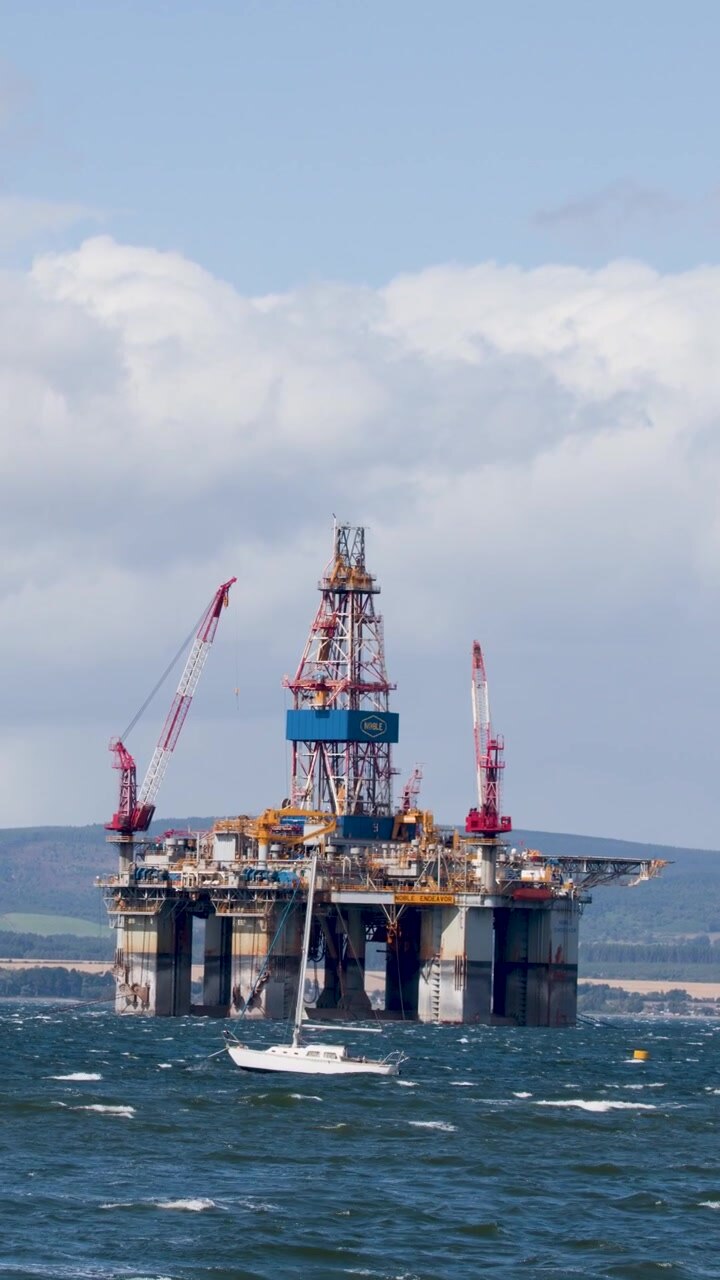 Offshore oil platform and passing yacht in daylight, wide shot, steady camera, partly cloudy sky