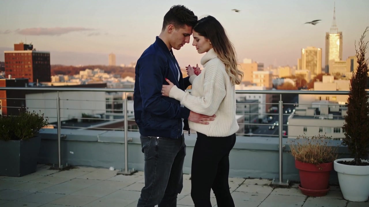 O pôr do sol aproximando o casal, que se abraça e toca as testas em um telhado perto de um guarda-corpo.