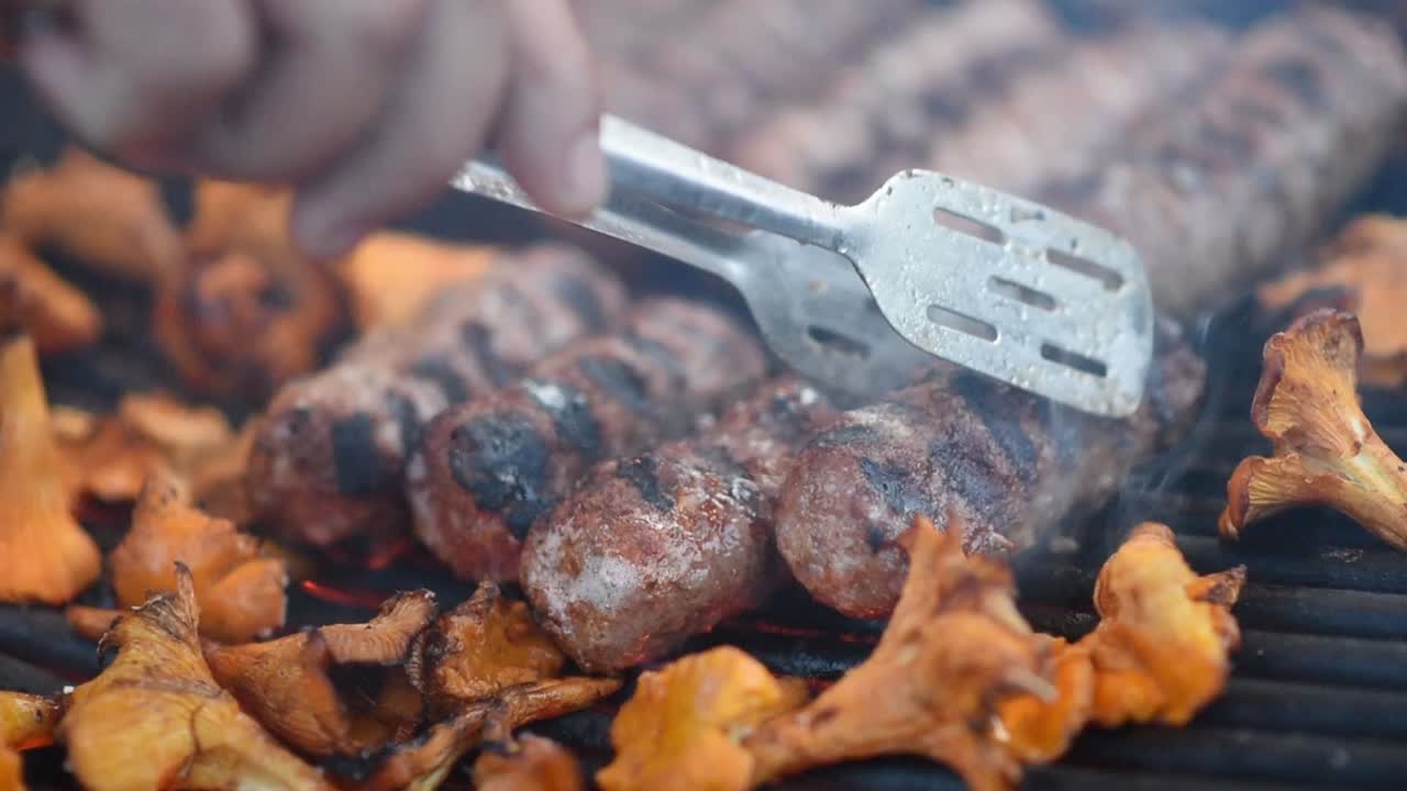 Man preparing sausage and mushrooms on the grill using tongs. Slow motion shot. Close up