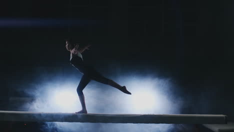 la niña realiza un truco en un tronco con luz de fondo y cámara lenta en ropa de gimnasia deportiva. humo y azul. saltar y girar en la viga de equilibrio