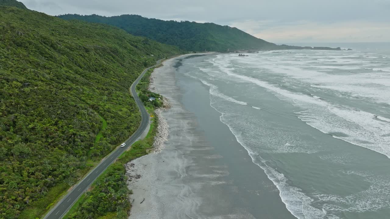 Aerial view following tourist campervan traveling along the wild, rugged and remote West Coast with rolling waves on the South Island of New Zealand Aotearoa
