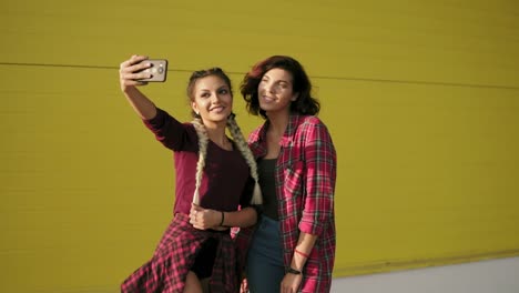 dos mejores amigas se divierten y se hacen selfies de pie junto a la pared amarilla. dos chicas hipster toman fotos de selfies con