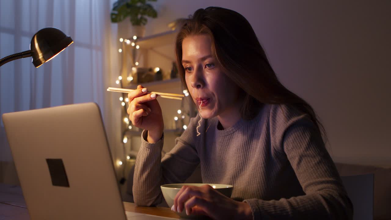Woman Eating Noodles While Working on Laptop at Night