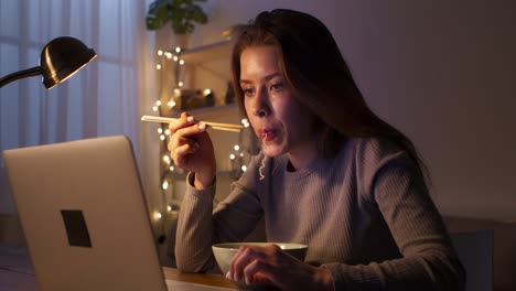 woman eating noodles while working on laptop at night