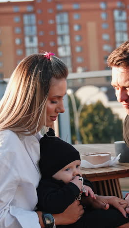 Happy smiling Caucasian couple sitting at the table outdoors. Man kisses his wife holding a baby in hands. Vertical video