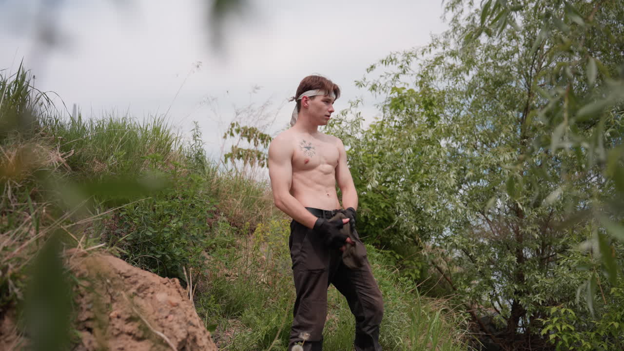 Fighter standing confidently on grassy riverbank, removing sleeveless top to reveal muscular upper body, surrounded by dense green foliage and earthy terrain under cloudy daylight in outdoor setting