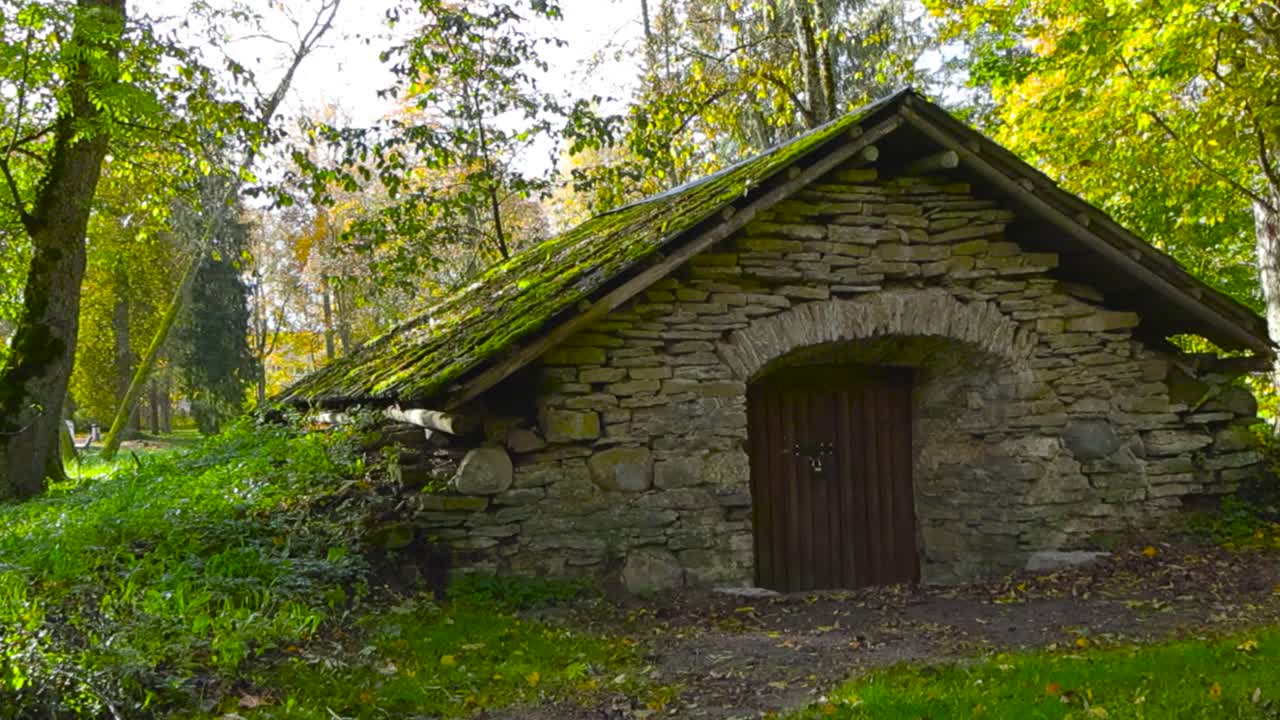 Old and vintage limestone building with a large wooden brown door in a autumn park during day time, what has a mossy stone roof. The building seems abandoned and maybe a cellar from the old times.