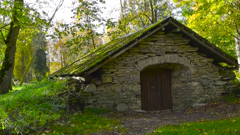 old and vintage limestone building with a large wooden brown door in a autumn park during day time, what has a mossy stone roof. the building seems abandoned and maybe a cellar from the old times.