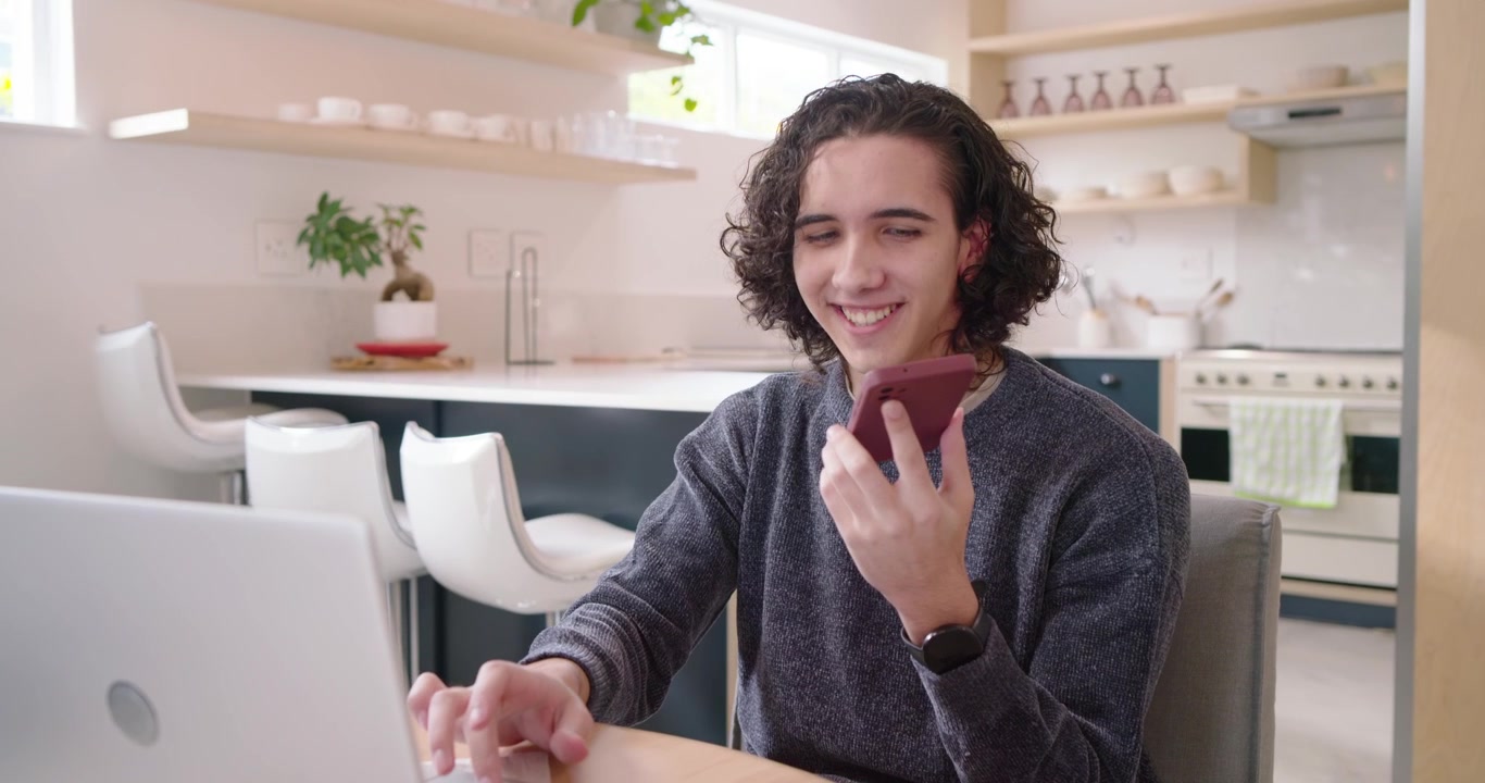 Uomo afroamericano ventenne che riceve una chiamata di lavoro, tiene in mano lo smartphone e digita sul laptop in cucina.
