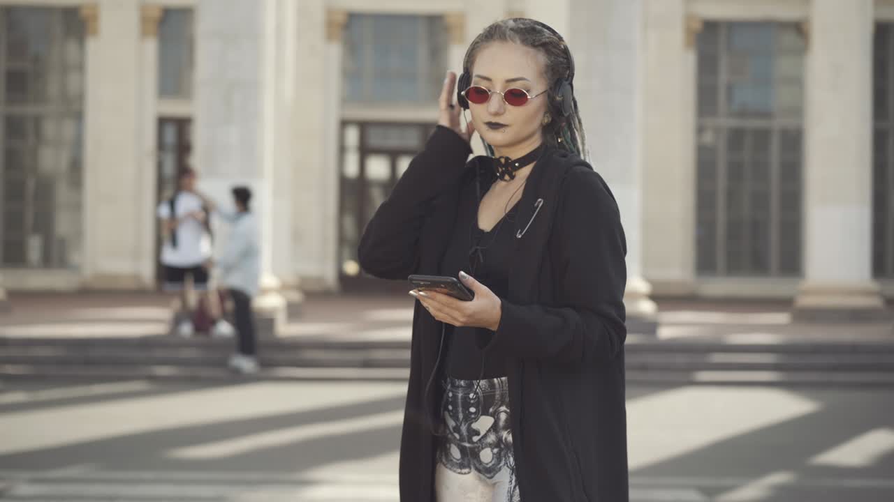 Carefree Caucasian hippie woman in sunglasses and headphones dancing on sunny city street with blurred men talking at background. Portrait of relaxed young goth enjoying music outdoors.