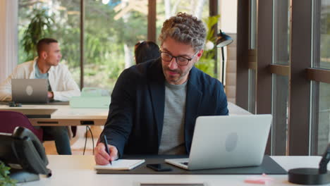 Mature-Businessman-Wearing-Glasses-Working-In-Office-Writing-In-Notebook