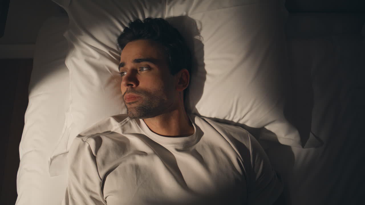 Guy lying dark bedroom at white bedding looking up closeup top view. Man on bed