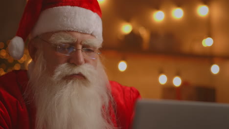 santa claus using laptop in living room. portrait of thoughtful santa claus typing with modern laptop in decorated house. high quality 4k footage