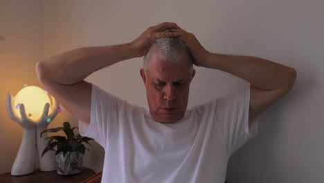 exasperated grey haired older caucasian man in white t-shirt holds head in frustration