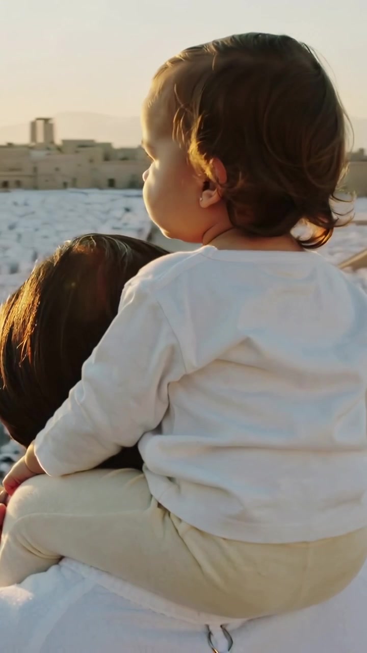 Video verticale: Uomo con maglietta chiara che tiene un bambino piccolo sulle spalle su un tetto, il sole che abbaglia, mano sulla coscia.