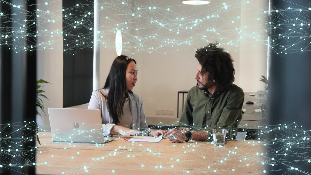 Woman at laptop greeting man approaching, reviewing docs as particles marking papers for business