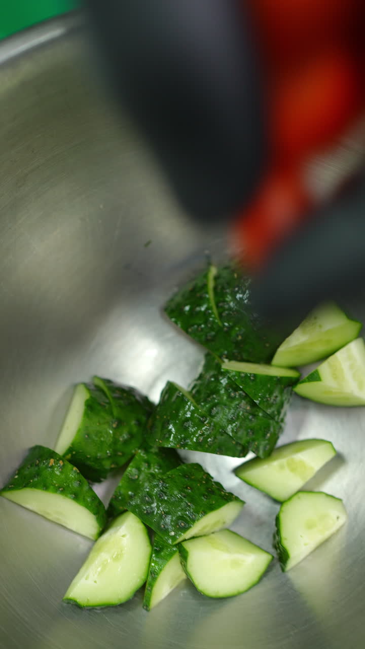 Vertical footage Hands in protective gloves chopping cherry tomatoes on green cutting surface, transferring freshly sliced produce into metal mixing bowl with cucumbers