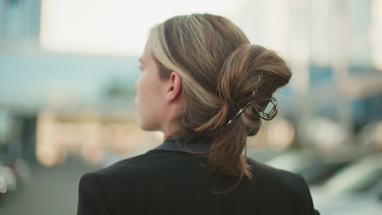Close up back view of woman with neatly clipped hair glancing left with blurred background of parked cars and modern glass building in city environment during bright workday morning in urban setting