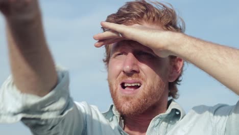 un primer plano de la cara de un hombre de cabello pelirrojo sombreando sus ojos mirando y apuntando al horizonte