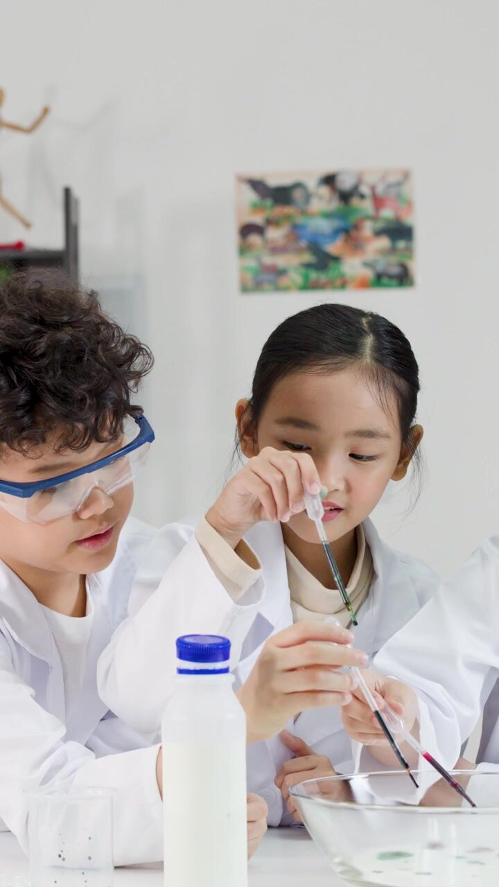 Asiatiske børn iført laboratoriekittel og sikkerhedsbriller bruger pipetter til et kemieksperiment.