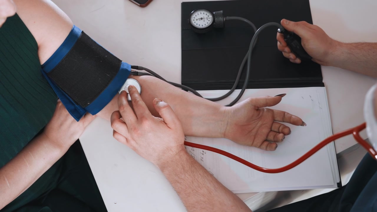 Donna di colore a cui viene misurata la pressione sanguigna mentre l'infermiera registra le letture su una tabella, bracciale sul braccio, manometro e tubi sul tavolo, mani che tengono penna e carta, visita domiciliare intima incentrata sulla prevenzione.
