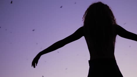 a woman raising her hands to the sky and nature, dancing under the purple night sky with bats flying high - slow motion