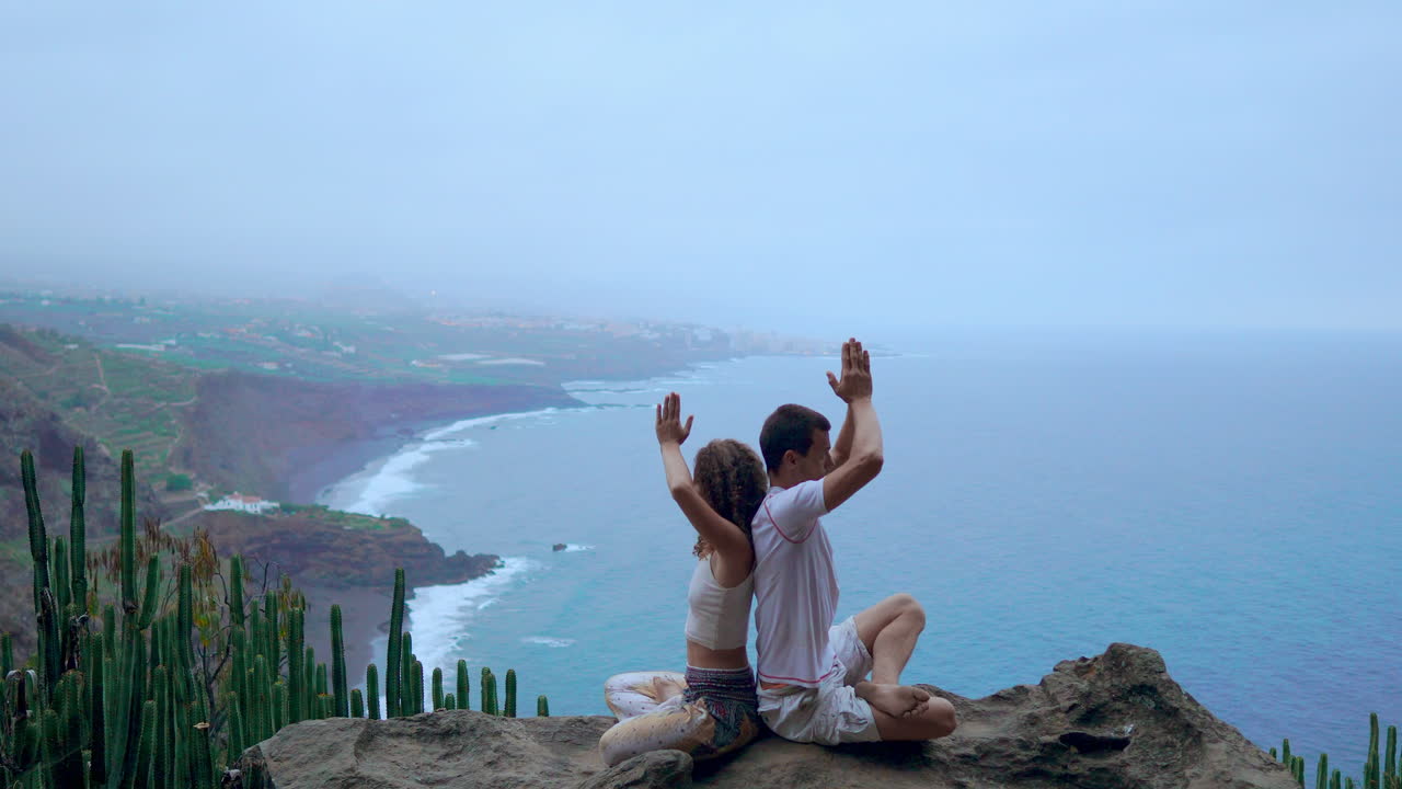 On a rock at the mountain's summit, a man and woman meditate and do yoga back to back, taking in the ocean's calming presence
