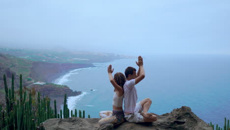 on a rock at the mountain's summit, a man and woman meditate and do yoga back to back, taking in the ocean's calming presence