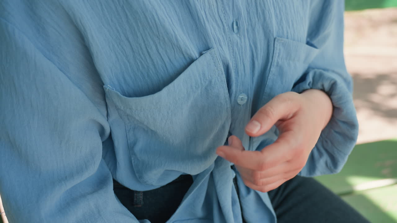 손이 블라우스 주머니와 매듭을 클로즈업해서 매만지는 모습이에요. 부드러운 파란색 천과 미묘하게 잡아당기는 동작이 보이고, 편안하게 매듭지은 넥타이를 맨 채 공원 벤치에 앉아 있어요. 기대감과 사적인 감정을 암시하는 섬세하고 초조한 몸짓입니다.