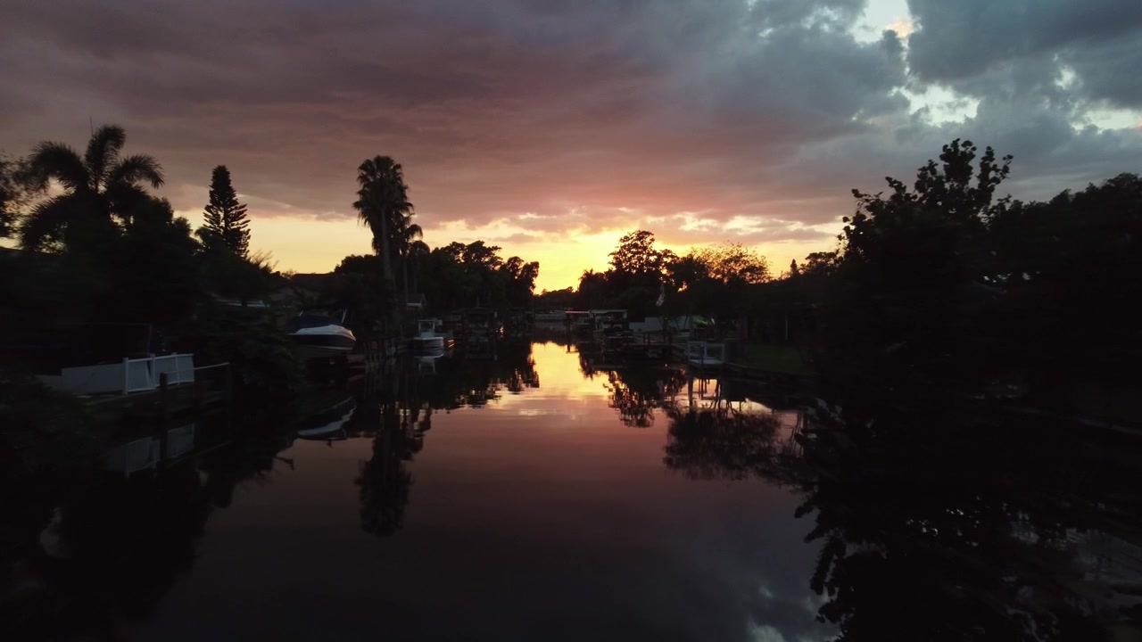 Pôr do sol em Palm Harbor, Flórida, ao longo de um rio de um condomínio de casas. A filmagem se move ao longo do rio mostrando os jardins das propriedades com pontões e barcos. Céus alaranjados à frente enquanto o sol se põe