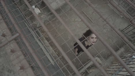 a gothic girl in black clothes with a scary smile stretches her arms up from the cage in which she is trapped