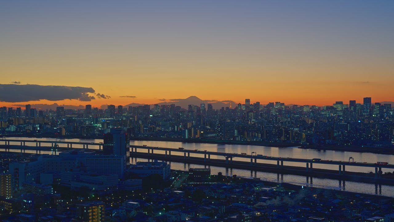Wide aerial view from Funabori Tower showing the glowing city lights at dusk. Mount Fuji is visible on the orange horizon behind the urban skyline.