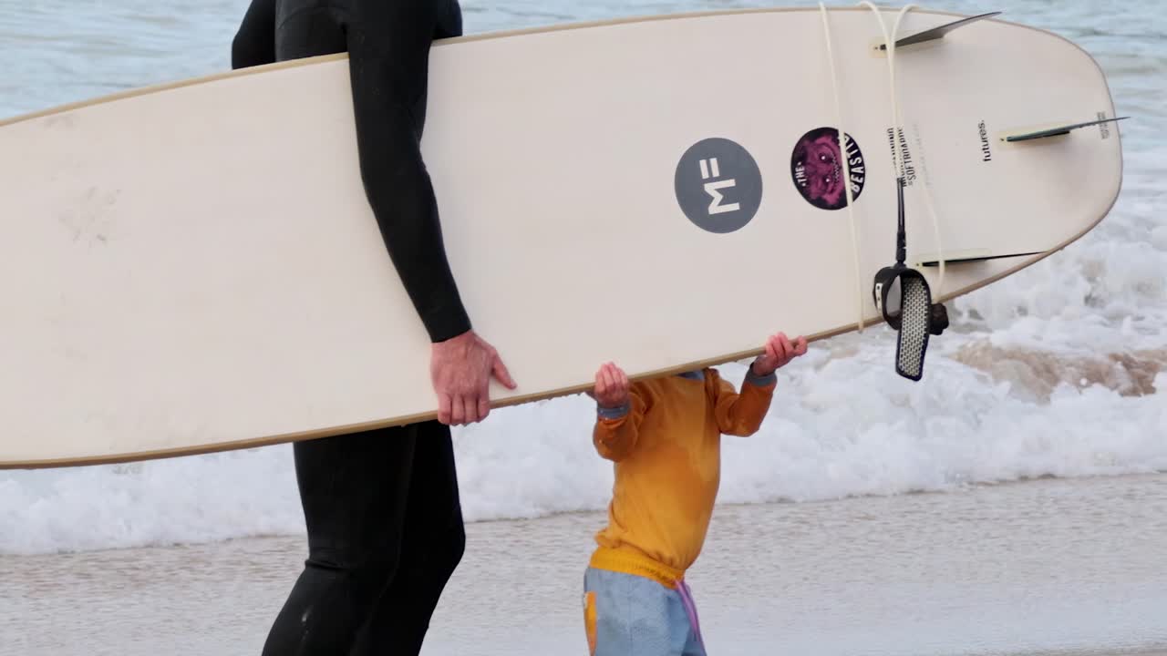 A child and adult walk along the beach carrying a surfboard near the ocean waves.