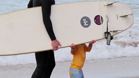 a child and adult walk along the beach carrying a surfboard near the ocean waves.